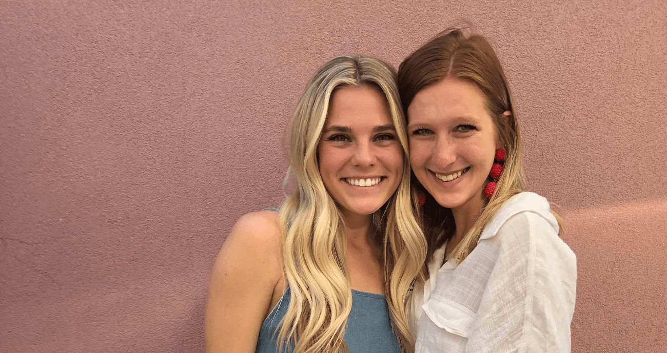 Hannah and Ashlyn are standing together by a pink wall. Both of them are smiling.