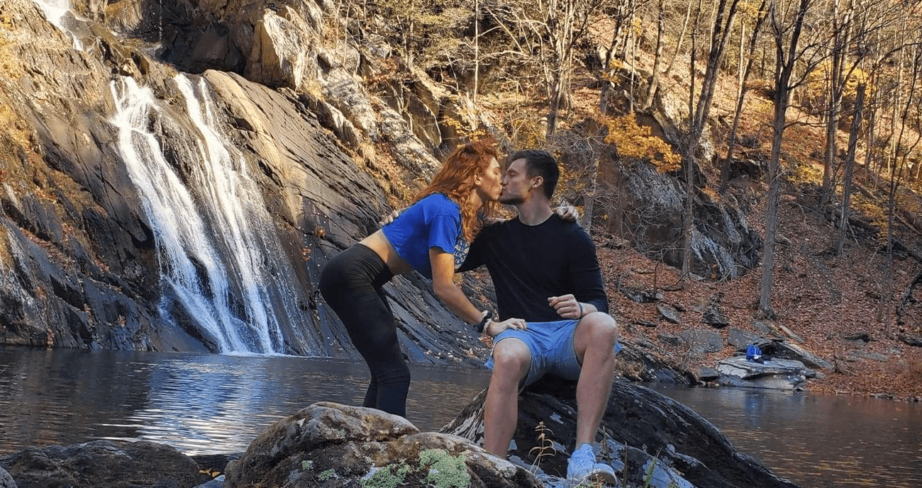 Paige and Shiloh share a kiss in front of a waterfall. Paige wears a blue top, while Shiloh is dressed in shorts.