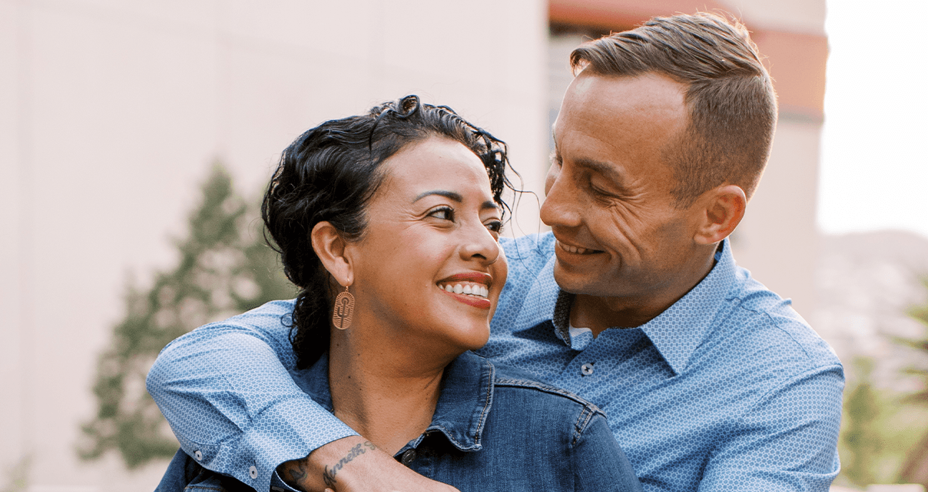 Matt and Iris are looking at each other. Matt has his arm around Iris's shoulder. She is wearing a blue denim jacket and Matt is wearing a blue shirt.