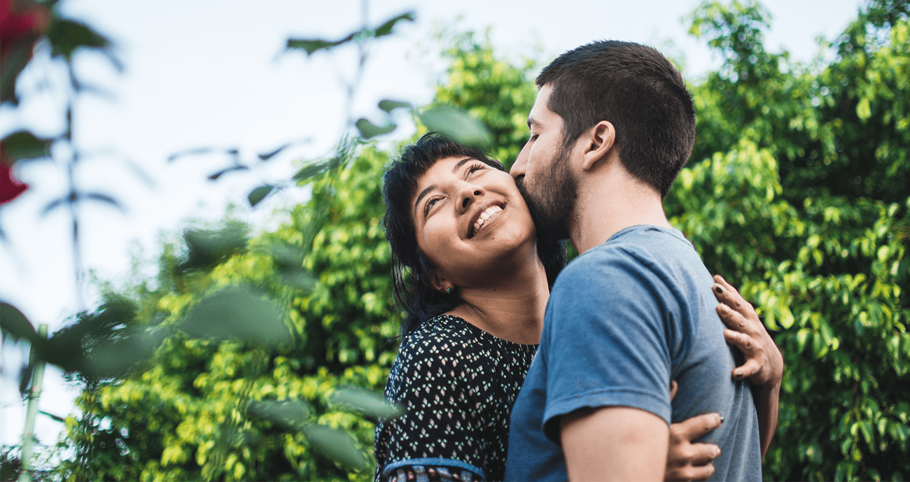 A couple embraces in a lush, green outdoor setting. The woman smiles with her eyes closed as the man kisses her on the cheek, conveying warmth and trust. The scene captures a moment of joy and emotional closeness, symbolizing transparency in a relationship.